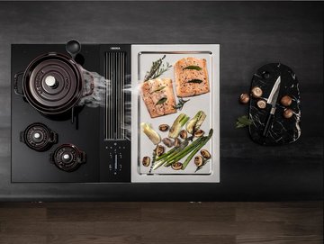 Roestvrijstalen grillplaat op een inductiekookplaat, met zalm en groenten erop. Ernaast staat een kookpot met rook die wordt opgezogen in een geïntegreerde downdraft motorkap.